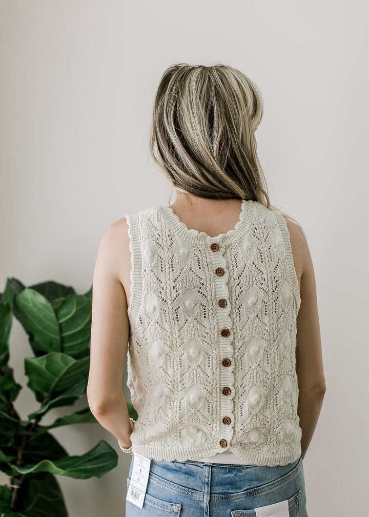 Back view of Model wearing a sleeveless cream knit sweater with brown buttons, and scalloping. 
