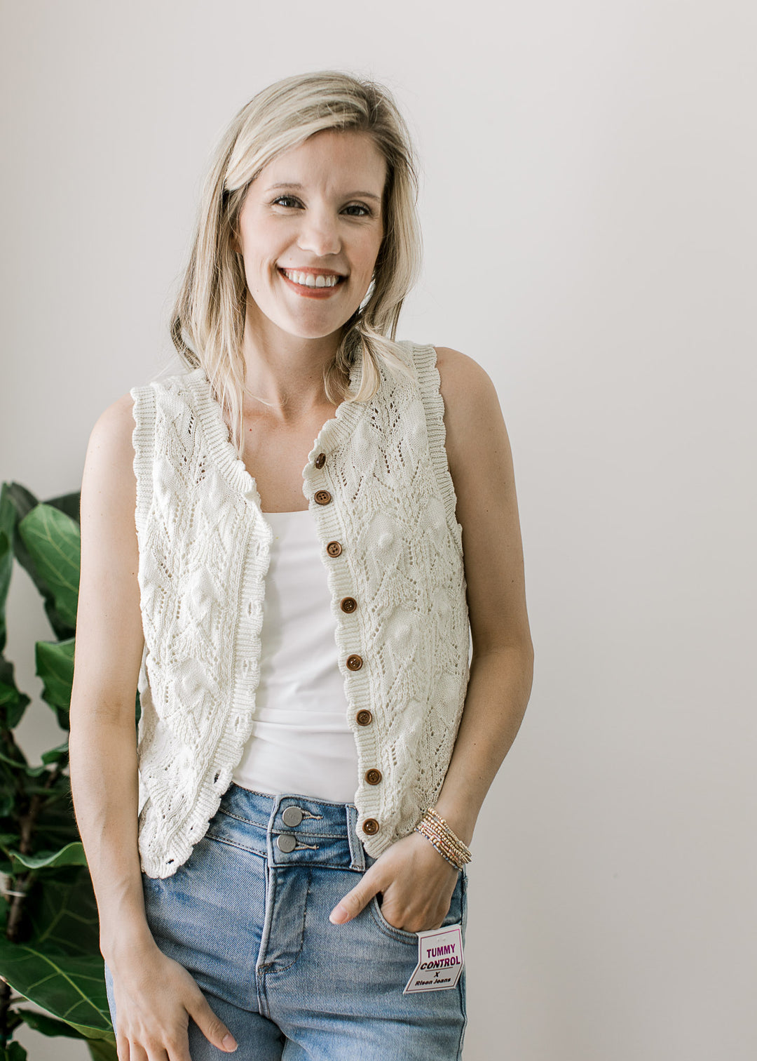 Model wearing a sleeveless cream knit sweater unbuttoned with a scalloping at cuff, neck and hem.