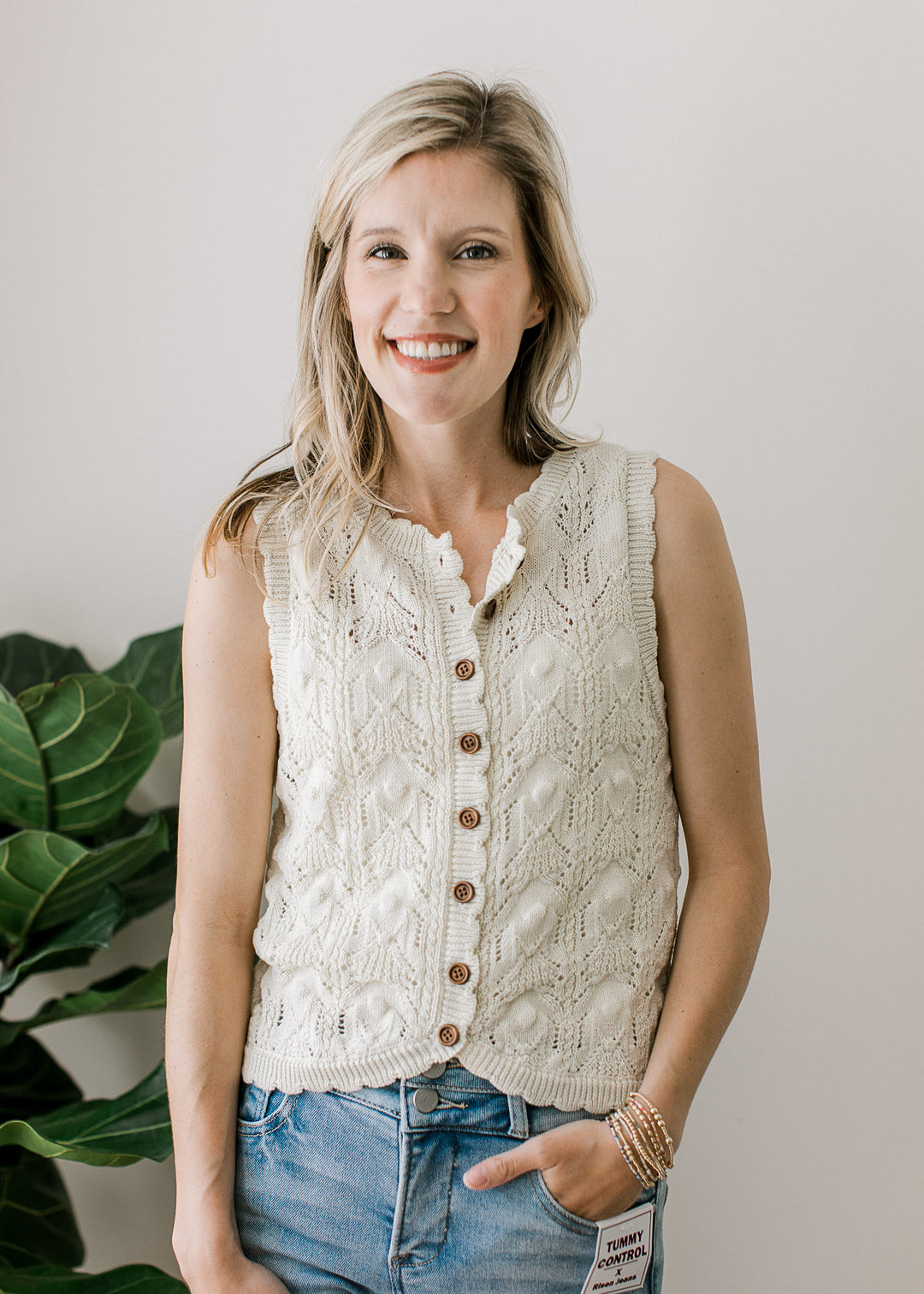Model wearing jeans and a cream knit sweater with brown buttons, scalloped neck, arm, and hem. 