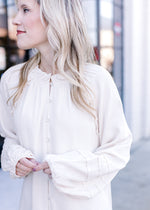 Close up of ruffle at neck and micro pleating on the sleeve of a long sleeve cream blouse.