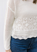 Close up of scalloped hem on a cream crochet sweater with long sleeves and extended shoulder.