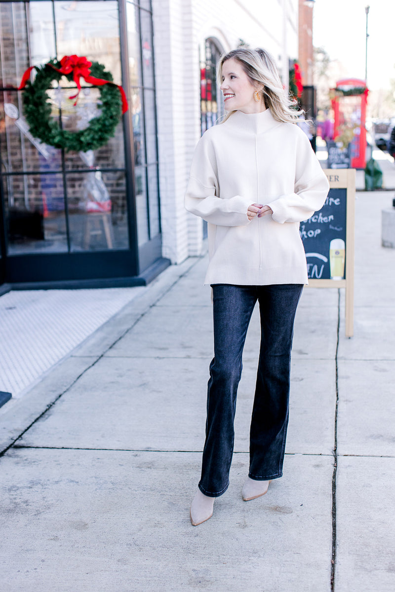 Model wearing jeans, booties and  a cream sweater with a mock neck and long sleeves.