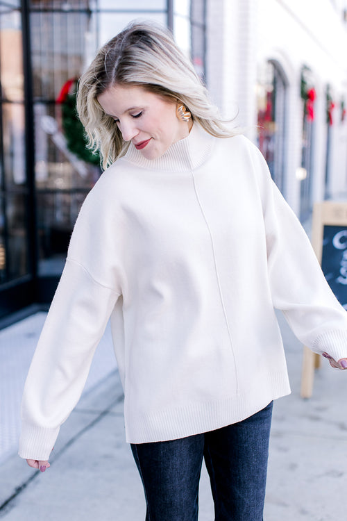 Model wearing a cream sweater with a mock neck, long sleeves and extended shoulder. 