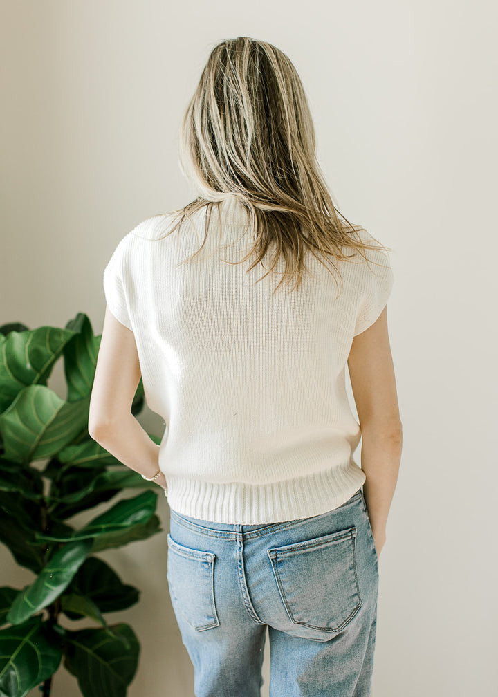 Back view of Model wearing a cream knit top with a ribbed mock neck and hem and short drop sleeves. 