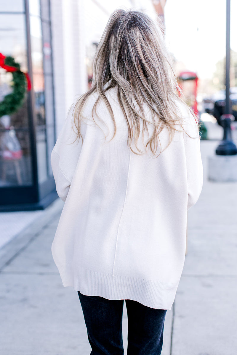 Back view of Model wearing a cream long sleeve sweater with a mock neck and extended shoulder. 