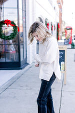 Side view of Model wearing a cream sweater with a mock neck, long sleeves and split sides.