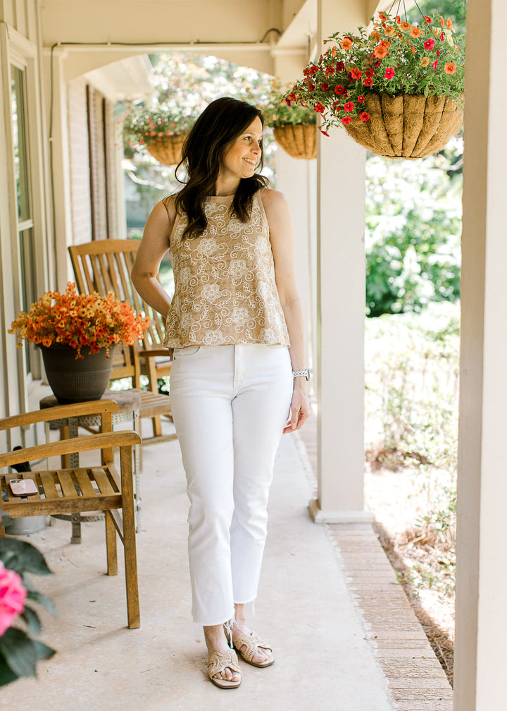 Model wearing white pants and a sleeveless camel top with cream floral stitching. 