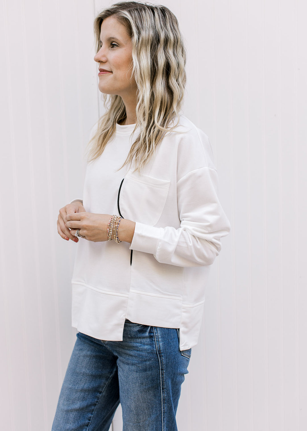 Model wearing a cream long sleeve top with black piping, patch pockets and square side cut out. 