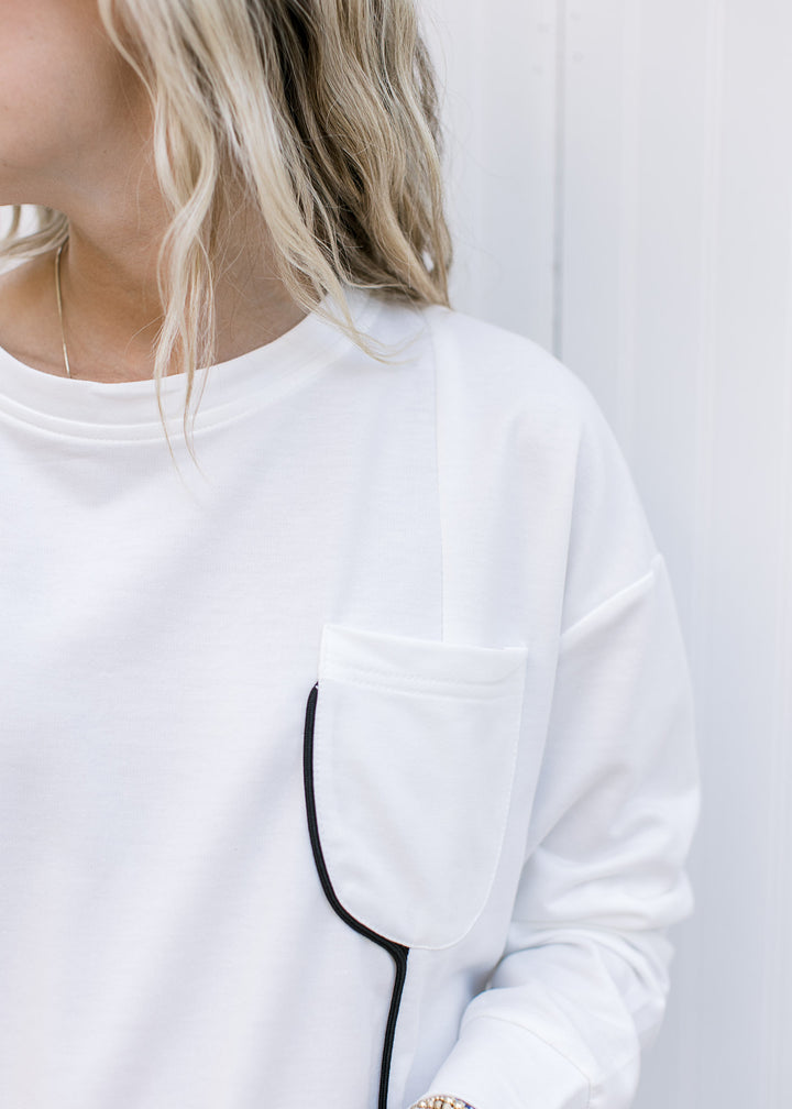 Close up of patch pocket and black piping on a cream top with a round neck and long sleeves. 