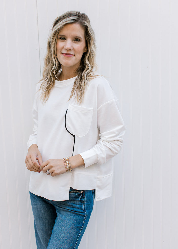 Model wearing jeans and a cream long sleeve top with black piping and patch pockets. 