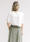Back view of a model wearing a cream v-neck top with a pleated shoulder detail and short sleeves. 
