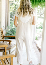 Back view of Model wearing a cream denim jumpsuit with wide legs and adjustable straps