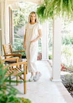 Model wearing wedges with a cream denim jumpsuit with adjustable straps and patch pockets.