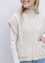 Model wearing a white top under a cream cable knit sweater with front pouch pockets.