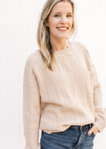Model wearing jeans with a cream cable knit sweater with a patch pocket and long sleeves. 