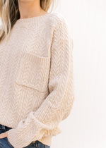 Close up of patch pocket and long sleeves on a cream cable knit sweater with a round neck. 