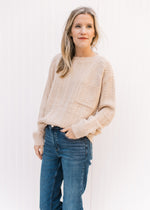 Model wearing a cream cable knit sweater with a patch pocket and long sleeves tucked into jeans.