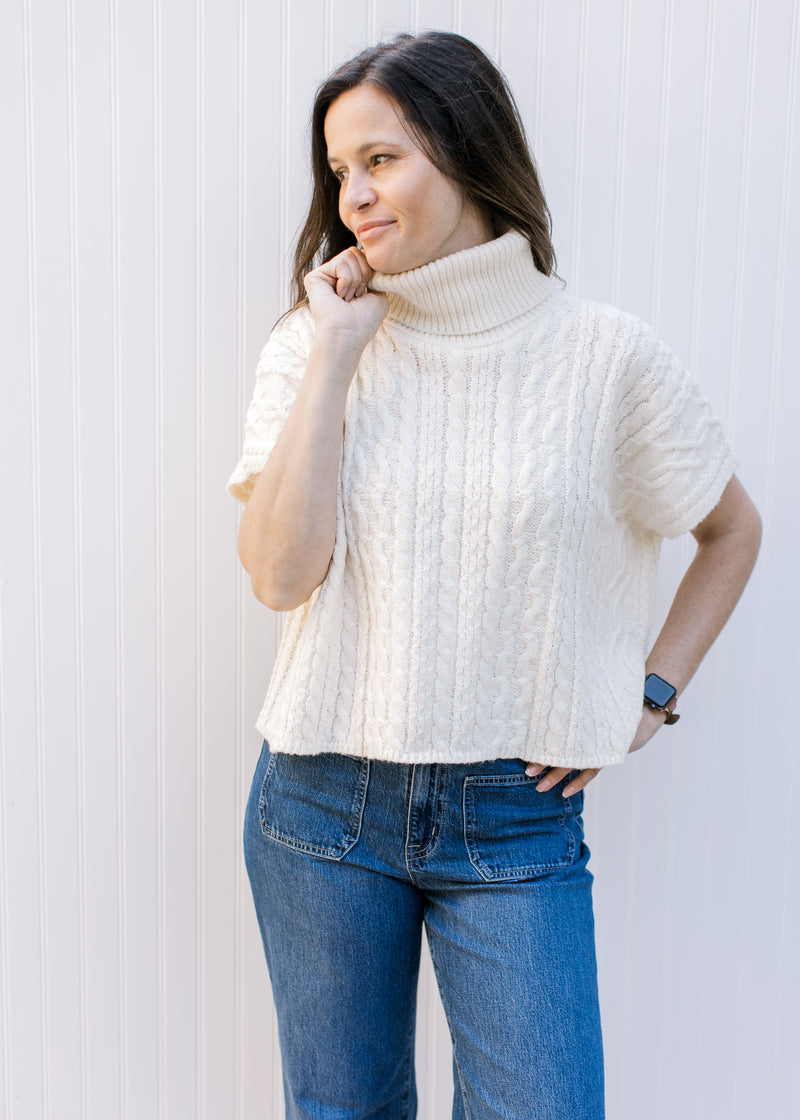 Model wearing a cream cable knit sweater with a slightly cropped fit, cowl neck and short sleeves. 