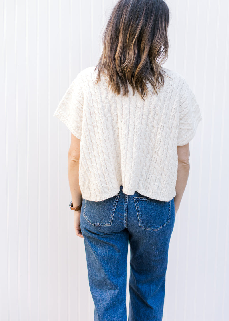 Back view of Model wearing a cream cable knit sweater with a cowl neck and short sleeves. 