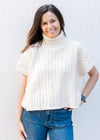 Model wearing jeans with a cream cable knit sweater with a cowl neck and short sleeves. 