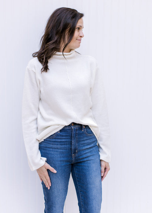 Model wearing a cream turtleneck sweater with long sleeves and a split hem at the cuff. 