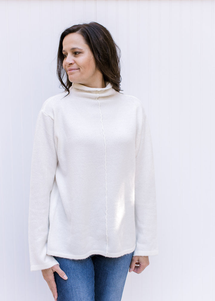 Model wearing jeans and a cream turtleneck sweater with long sleeves and a split hem at the cuff. 
