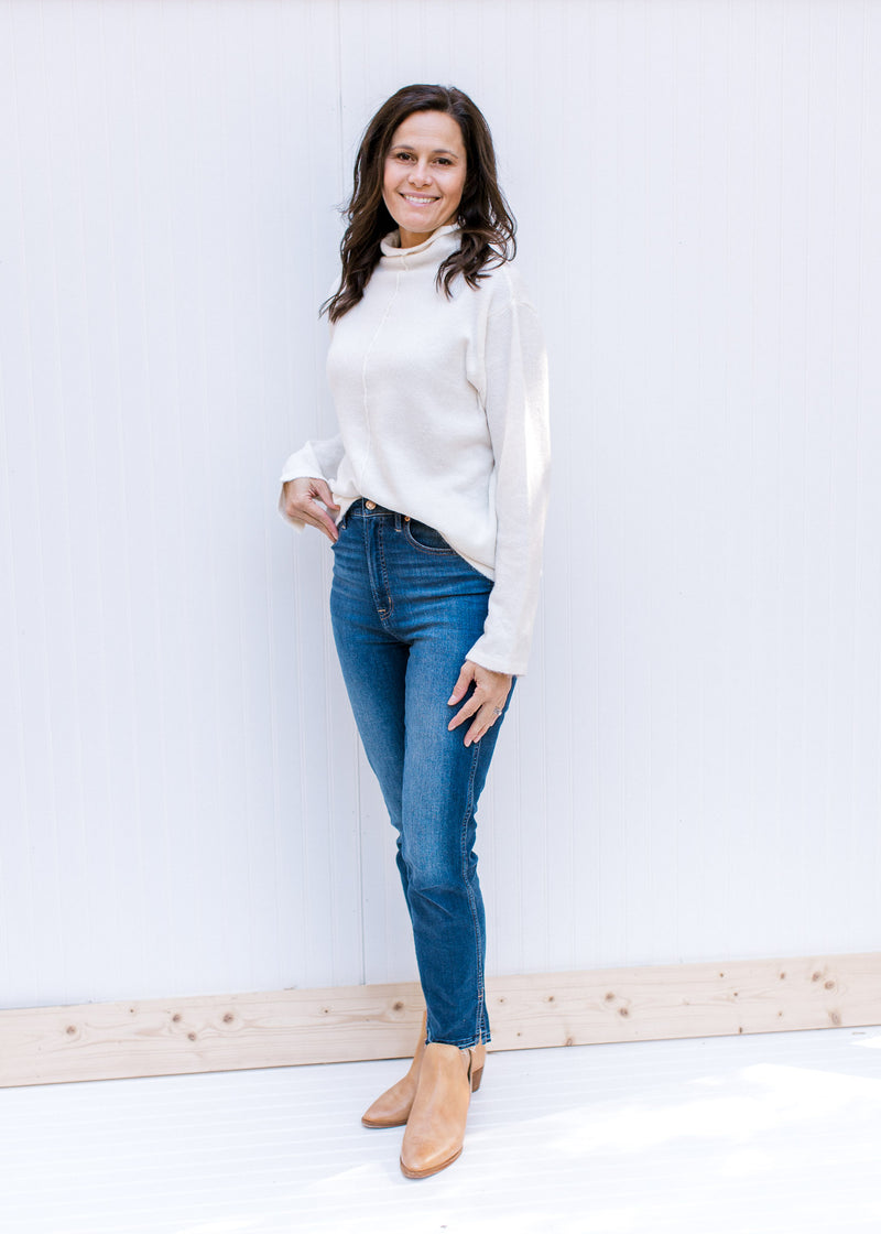 Model wearing jeans, mules and a cream turtleneck sweater with long sleeves and a split cuff hem. 