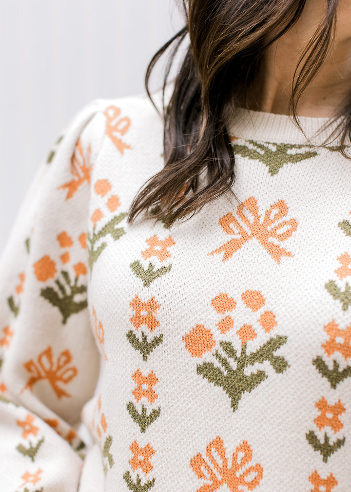 Close up of pleated shoulder on a long sleeve cream sweater with orange and green floral. 