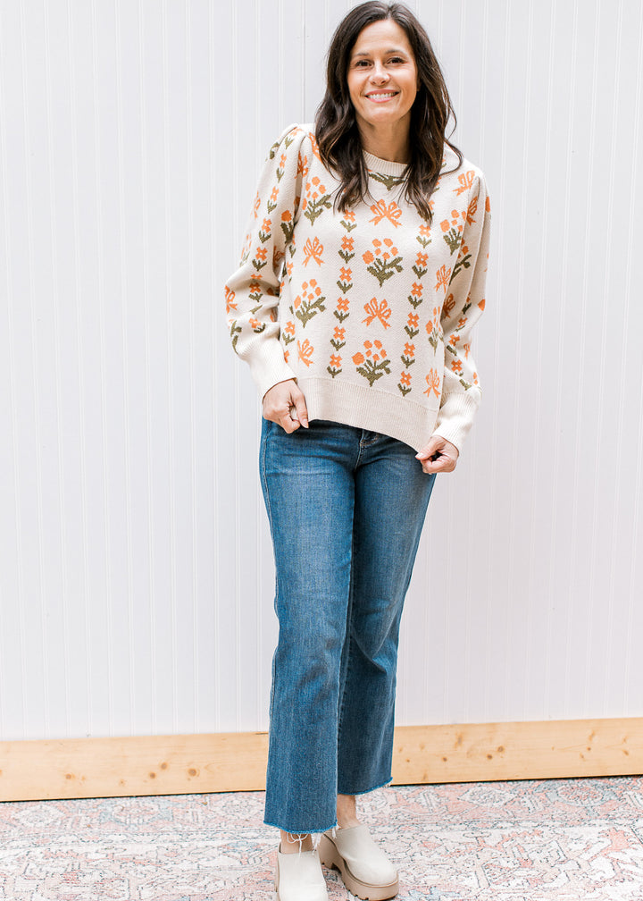 Model wearing jeans and a cream sweater with orange and green floral, round neck and long sleeves. 
