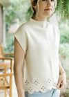 Model wearing a cream short sleeve sweater with a mock neck and eyelets at the scalloped hem.