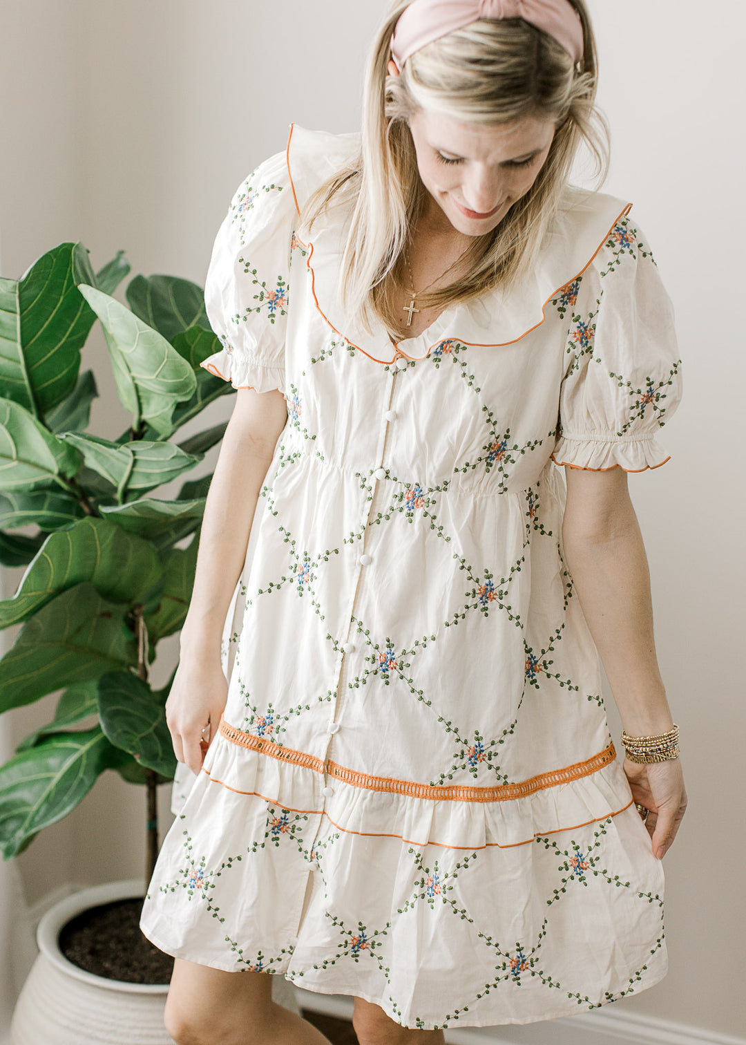 Model wearing a cream dress with embroidered lattice pattern and rust and blue floral accents.