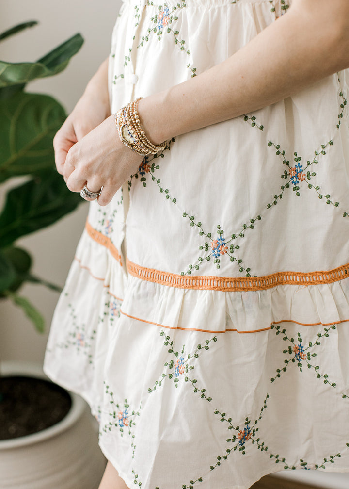 Close up of embroidered lattice pattern and rust and blue floral accents on a cream dress.