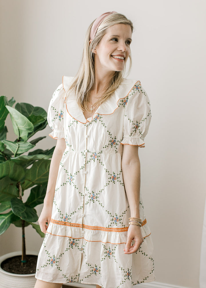 Model wearing a cream above the knee dress with embroidered floral, v-neck and short sleeves. 