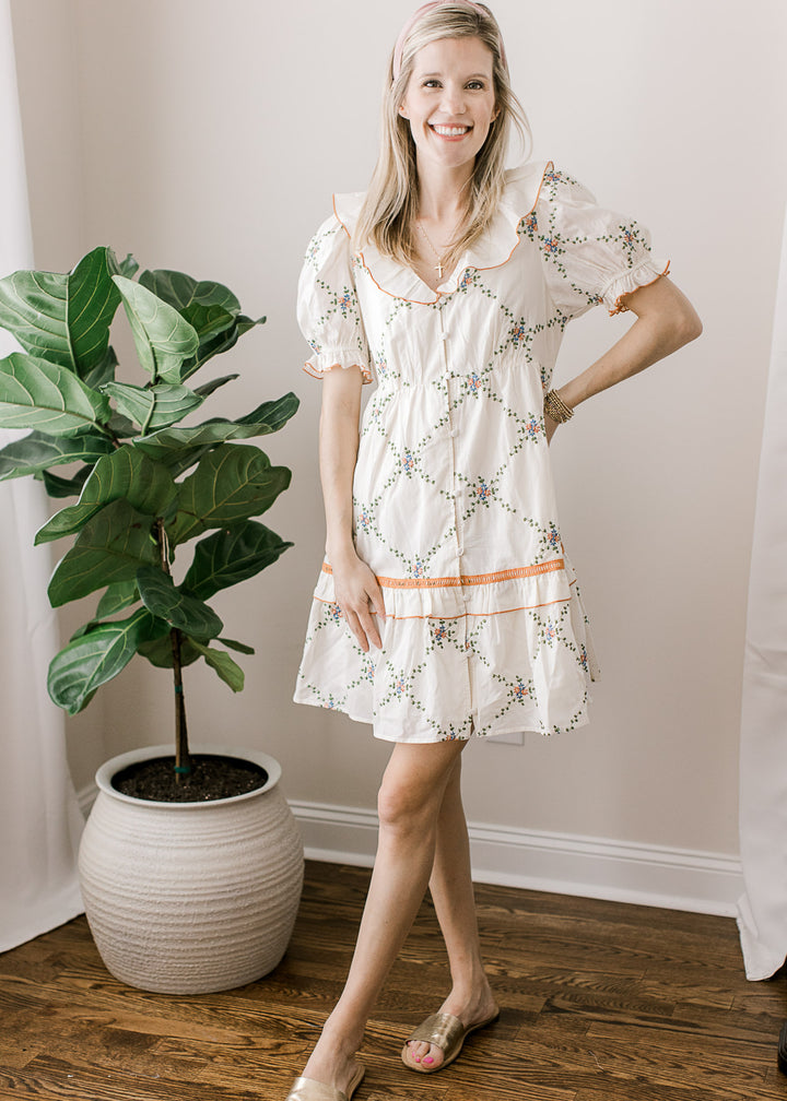 Model wearing sandals and a cream dress with embroidered lattice pattern and rust and blue floral.