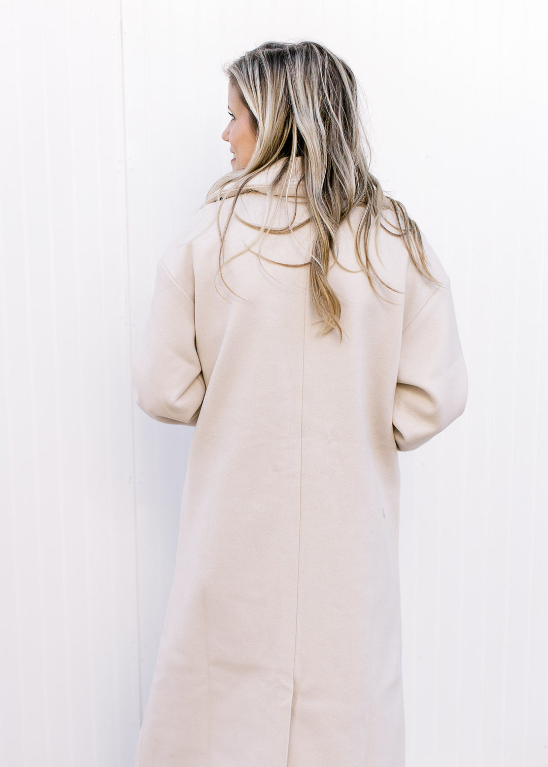 Back view of Model wearing a cream long line jacket with split back, long sleeves and a collar. 