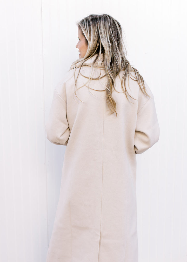 Back view of Model wearing a cream long line jacket with split back, long sleeves and a collar. 