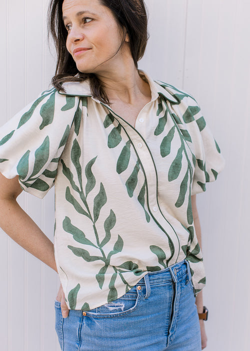 Model wearing a cream top with olive print, bubble short sleeves and button closure. 