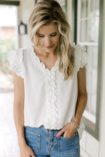 Model wearing a white top with ruffle cap sleeves, ruffle at v-neck and down placket. 