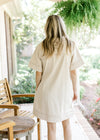 Back view of Model wearing a cream denim dress with contrast stitching ,v-neck and short sleeves.