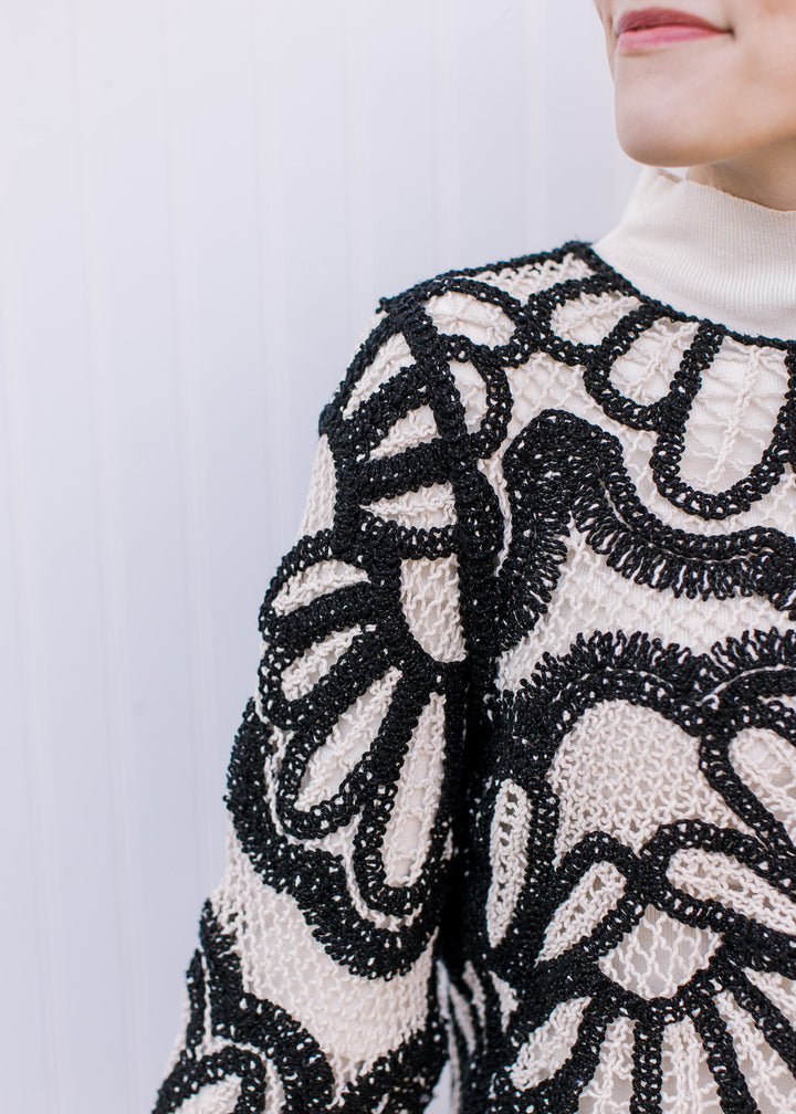 Close up of black graphic design on a cream open knit sweater with long sleeves and round neck. 