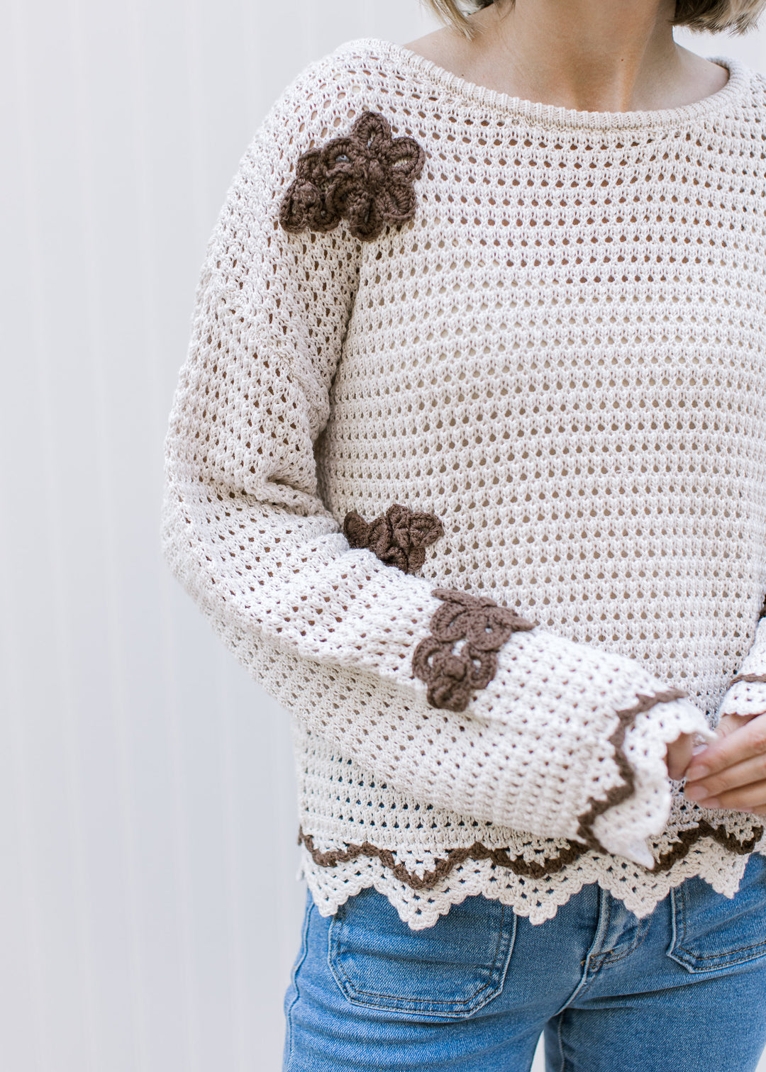 Model wearing a taupe open knit sweater with brown flowers, long sleeves and scalloped detailing.
