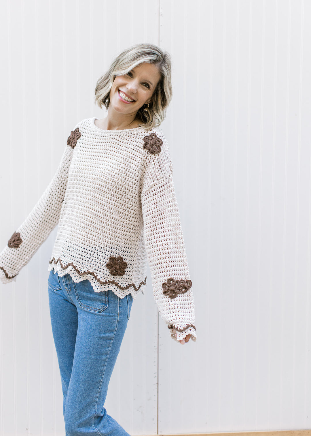 Model wearing jeans and a taupe open knit sweater with brown crochet flowers and scalloping.
