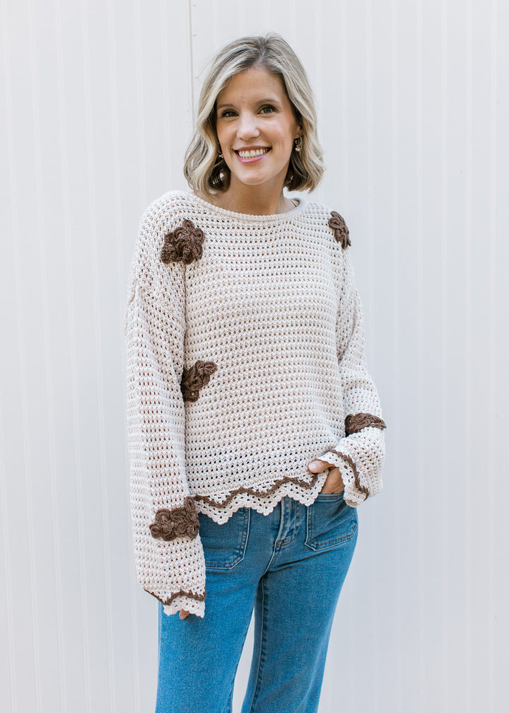 Model wearing a taupe sweater with brown crochet flowers, long sleeves and scalloped detailing.