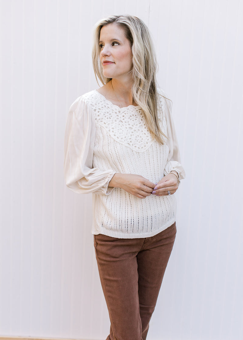 Model wearing a cream top with crochet front, pleated shoulders and 3/4 sleeves with elastic cuff. 