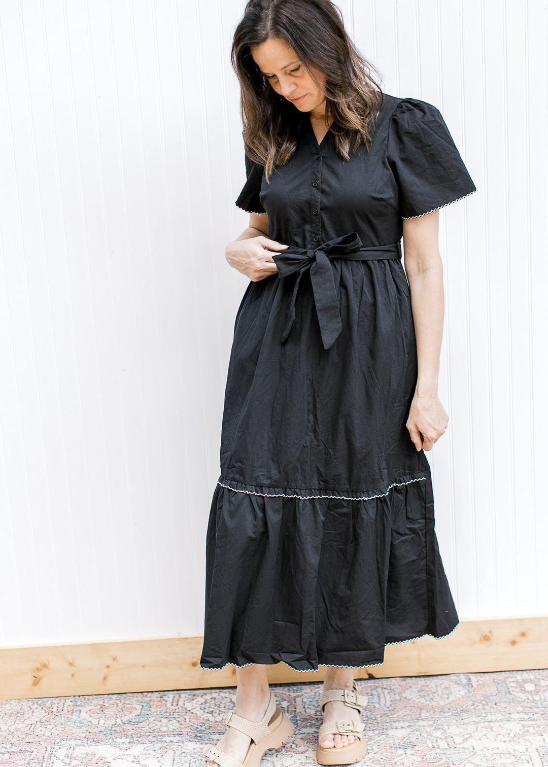 Woman wearing a black midi with puff sleeves, v-neck, buttons and a tie waist. 