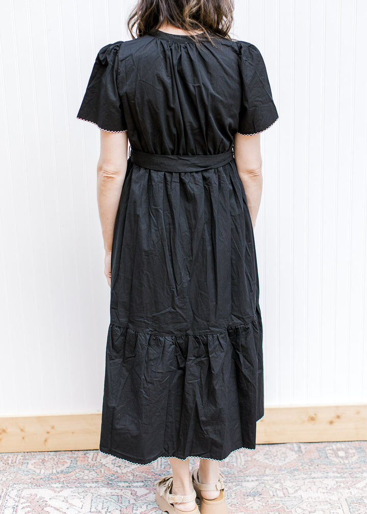 Back view of model wearing a black midi with short sleeves, tie waist and pleated shoulders. 