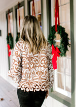 Back view of Model wearing a cream and reddish-brown lacework top with a lining and long sleeves.