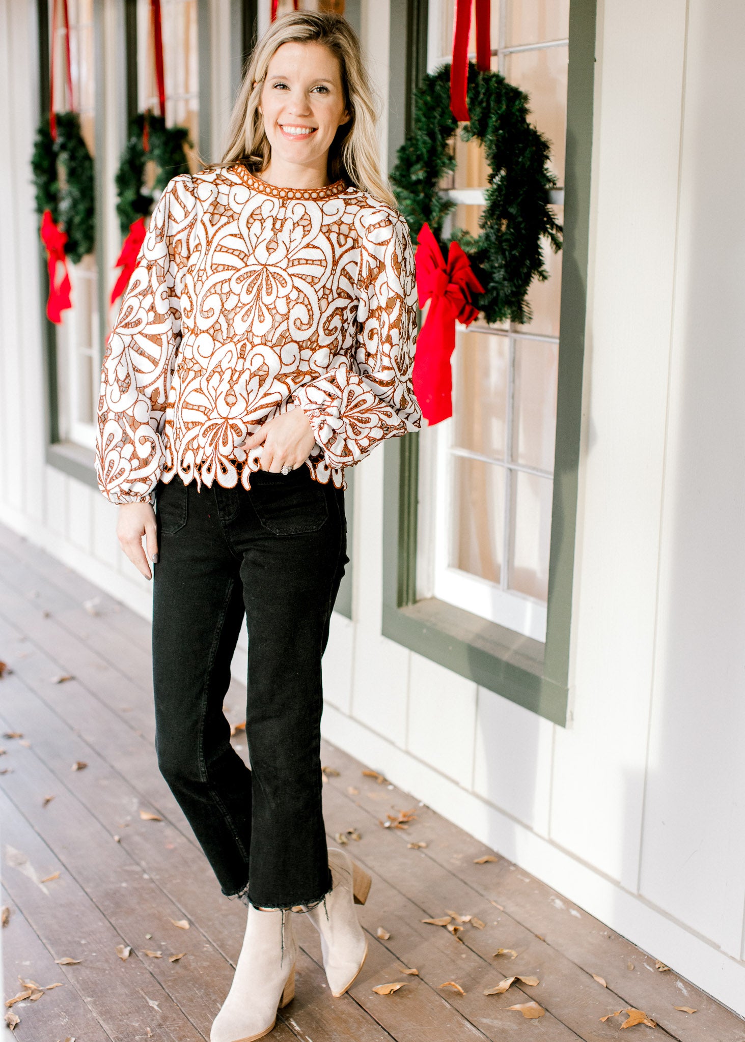 Model wearing black jeans and booties with a cream and reddish-brown lacework top with long sleeves.