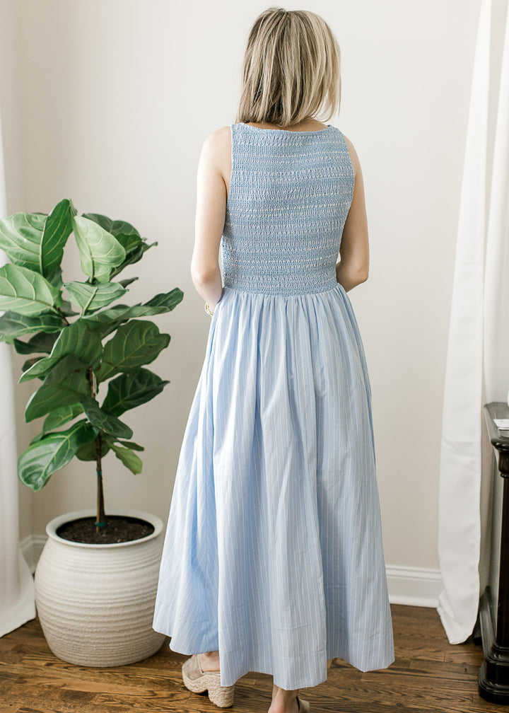 Back view of Model wearing a soft blue sleeveless maxi with white pinstripes and smocked bodice. 
