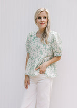 Model wearing white jeans with a sage babydoll top with cream daisies with puff short sleeves.